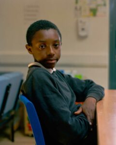 The picture is a portrait of a black young boy seated at his desk.