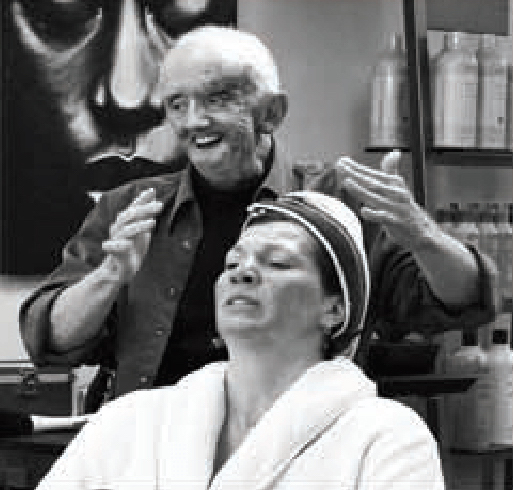 The black and white picture of a woman seated and her hairdresser moving his hands around her head.
