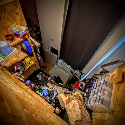 This is a high-angle, distorted shot looking down into a cluttered and narrow space, likely a room or a section of a room. The perspective creates a sense of being squeezed in or looking into a very cramped area. On the left, there's a wooden structure made of light-colored chipboard, possibly shelving or part of a built-in unit. It holds various items: cans of paint, what looks like a small wooden bowl, and other indistinguishable objects on shelves. The floor of the space is barely visible, but there's a jumble of items scattered across it. A light-colored electric guitar with a black pickguard is prominently featured in the center, lying amidst other objects. Near the guitar, there are small containers, tools, and what appears to be a piece of wood with a rough, natural edge, possibly a log or a decorative piece. To the right, there's a dark curtain hanging, and beyond it, a wall with some electrical outlets or switches. Further to the right, another wooden structure, similar to the one on the left, holds clear plastic containers with blue lids, stacked neatly. A clamp or vise is attached to this structure, suggesting a workspace. The overall impression is one of disarray and a lack of open space, with items piled and scattered, making it feel like a workshop or storage area that is very full.